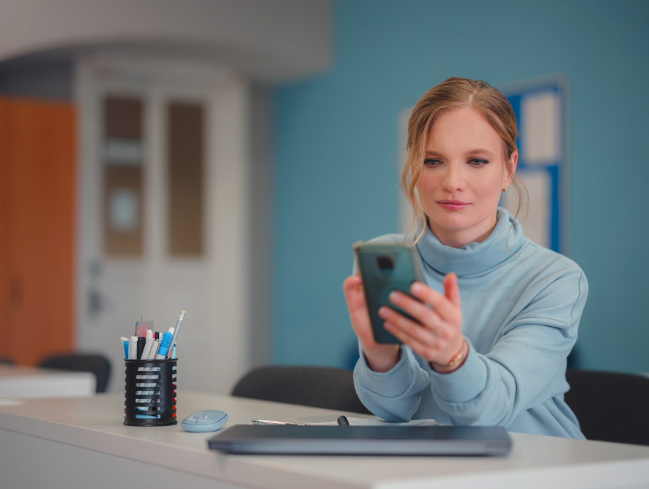 Woman sitting at a desk looking at a smartphone, with a tablet, mouse, and office supplies nearby.
